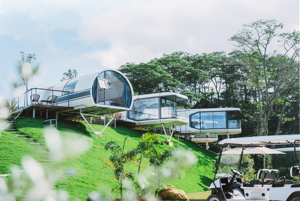 Futuristic capsule houses positioned on elevated platforms in a lush green landscape, offering panoramic views of the surrounding nature.