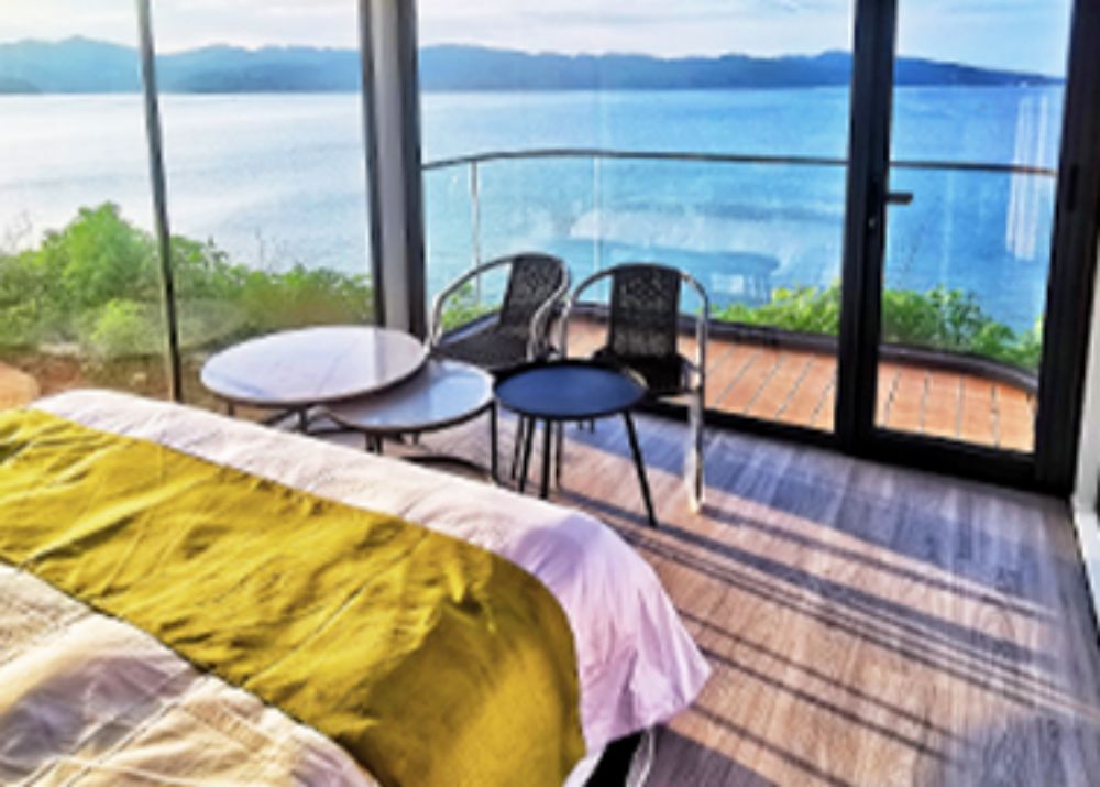 Capsule house interior featuring a bedroom with expansive windows overlooking a lake, cozy bedding, and balcony chairs
