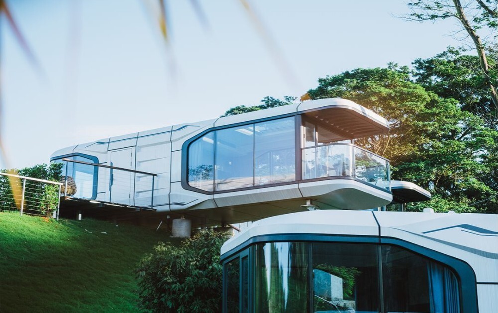 Luxurious capsule house on raised supports, featuring extensive glass walls and a panoramic deck, nestled in a lush, green setting.