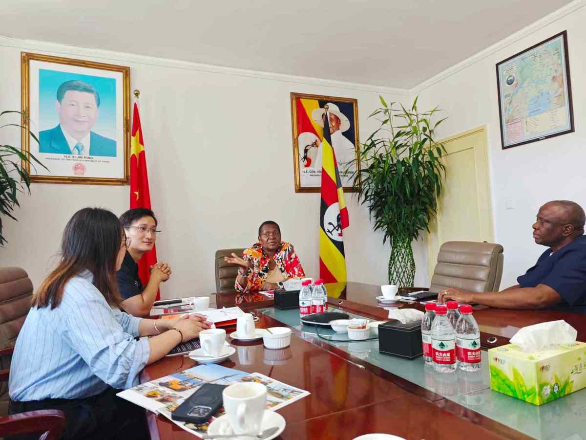 An overview shot of a comprehensive business meeting between ZW Group and the Ugandan Embassy delegation in Beijing. Ambassador Oliver Wonekha gestures while discussing infrastructure, with corporate brochures and branded water bottles on the table.